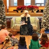 A woman dressed as a snow queen, seated between two decorated white Christmas trees, reads to a group of children on a library floor. 