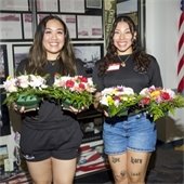 Two women stand side by side each holding two vases of red and pink flowers.