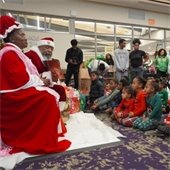 Santa and Mrs. Claus sit in a library, surrounded by joyful children in festive pajamas. A Christmas tree and gift bags add to the holiday spirit.
