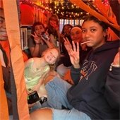 A group of smiling people in a decorated space with orange streamers and string lights. They pose with peace signs, conveying a cheerful, festive mood.