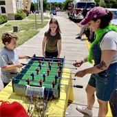 Two children playing foosball on a yellow table outside with a Community Engagement Staff Member