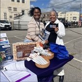 Two smiling people stand at an outdoor table with library sign-up materials under a blue sky. Flyers and a blue bag are on the purple-covered table.