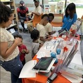 Four children and one adult stand at a robotics table coloring sheets of paper.