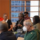 A diverse group of adults sit in rows of chairs at a conference. A woman in the center holds a microphone, speaking. 