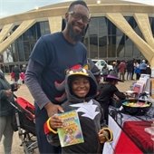 Father and child at an outdoor event, with the child wearing a dragon costume and holding a book. Smiling, they stand by a table with candy, surrounded by festive attendees. 