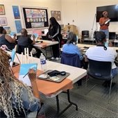 A few adults sit at tables in an open room painting blue skies on canvases. Camera shot is taken from behind.