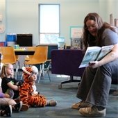 A woman reads a book to a group of young children, some seated and one in a tiger costume. The setting is a brightly lit classroom. 