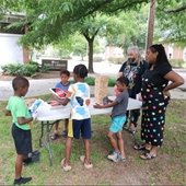 Four children and two adults stand around a folding table outside of the Blyden Branch Library. The children are playing uno at the tabkle with very large cards.
