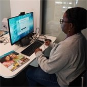 A person sits at a desk using a computer, focused on the screen displaying a login page. Brochures and informational materials are scattered on the desk.