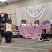 A woman reads a book to young children sitting on the floor in a cozy library setting. A table with books is covered with a pink cloth and festive decorations hang above.
