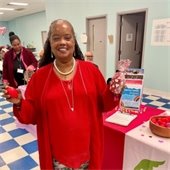 A woman in a bright red outfit stands smiling in a community room. She holds a small red ball and a gift bag. The atmosphere is festive and welcoming.