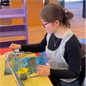 A person in glasses and a plastic apron paints a colorful canvas at a table. They look focused, with art supplies and a paint palette nearby.