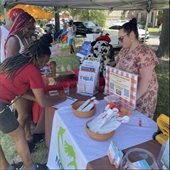 People engage at a community fair booth under a canopy. A woman in a floral dress interacts with two visitors beside a basket of flyers and giveaways.