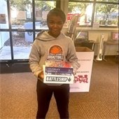 A person in a gray basketball hoodie holding board games like "Battleship" stands indoors. 