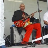 Man with red pants sits on an amp on a stage playing a red guitar.