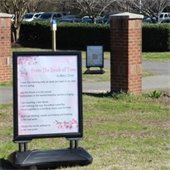 A park scene featuring a framed poem near two brick posts, a background of leafless trees, and a distant parking lot with cars. 