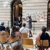 Two women stand in front of a seated crowd performing a Storytime. One woman is holding an open picture book, pointing to it.