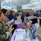 Outdoor community event with four women at an information booth. They discuss papers on a purple and white table, surrounded by brochures and snacks.