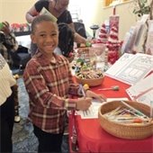 Smiling boy in a plaid shirt colors at a festive table with crafts, surrounded by a joyful group. Red decorations create a cheerful holiday atmosphere.