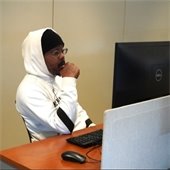 A person in a white hoodie and black hat sits at a desk, focused on a computer screen.