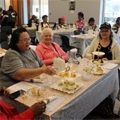 A diverse group of women enjoys a social gathering in a bright room. They sit at tables with lace tablecloths, chatting and laughing over tea and snacks.