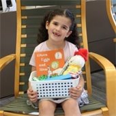 A young girl with curly hair sits on a wooden chair in a library, smiling and holding a basket containing the book "Green Eggs and Ham" and plush toys.