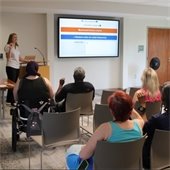 Instructor stands at the front of the room next to the tv doing sign language. The class of adults sit in chairs in front of her, signing back.