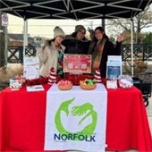 Three people smiling under a black canopy with a red-covered table. The table features holiday decorations, bowls of candy, and a "City of Norfolk Library" banner. 