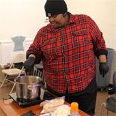 A person in a red plaid shirt and black beanie cooks over a pot on a portable stove in a classroom. Nearby are cooking ingredients on a table.