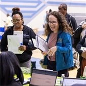 A group of people, smiling and holding documents, engage with individuals seated at a table with laptops. The atmosphere is lively and collaborative.