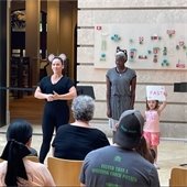 A woman with a mouse ears headband, another woman with cat ears, and a little girl holding a sign stating "fast" stand in front of a sitting audience in an open room