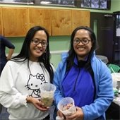 Two smiling individuals hold bowls of ice cream and toppings in a brightly lit room. A table with more toppings is in the background, along with another person preparing food.