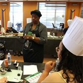 A woman leads a cooking class, demonstrating with spices. Participants in white chef hats watch attentively at a table with cooking ingredients.