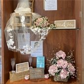A wedding display with a wooden chair draped in lace, a pink floral bouquet, a "Mr & Mrs" sign, champagne flutes, wedding book, and pink hydrangeas.