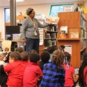 A woman reads a book to a group of young children seated on the floor in a library. Shelves with books and large windows are visible in the background.