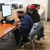 Two people focus on a computer in a room with cubicles; one wears a mask and the other a red cap, while another person sits behind them. Casual, focused atmosphere.