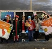 A group of six people in festive attire stand in front of a van at dusk. Two hold signs reading "The Snowy Day," suggesting a holiday theme.