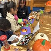 Children engaged in a craft activity, surrounded by colorful toys, pumpkins, and art supplies on a gold tablecloth. The joyful scene conveys creativity.