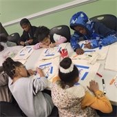 Children engaged in an art activity at a table with markers and drawings, focused and creative. The room has green walls and a relaxed atmosphere.