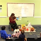 A person reads a Thanksgiving story to children seated on the floor in a library. A decorated board says "Happy Thanksgiving," and books and pumpkins are arranged nearby. 