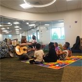 A man plays guitar to a group of children. They are all sitting on a colorful floor mat.