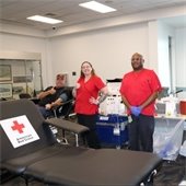 Two people in red shirts stand inside a room with Red Cross medical equipment. A man donating blood lays next to them giving the camera a thumbs up.
